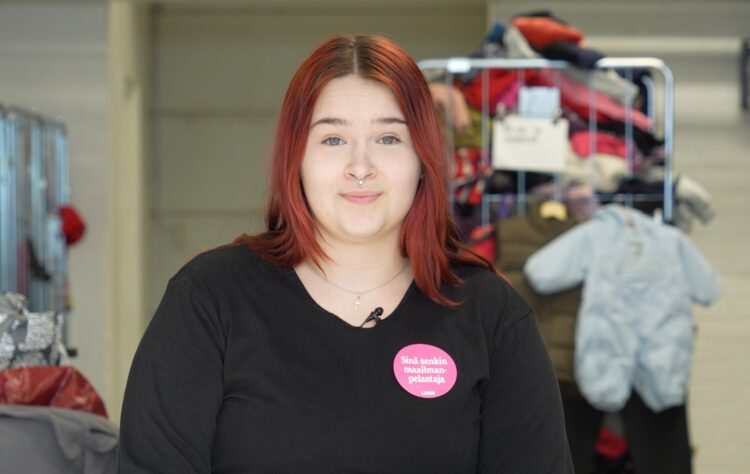 Nainen seisoo sisätiloissa, yllään musta paita, jossa on pinkki merkki. Taustalla on vaatteita rullakossa.