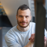Hymyilevä mies katsoo kameraan sisätiloissa. Hänellä on lyhyet hiukset ja parta, ja hän on pukeutunut vaaleansiniseen puseroon.
