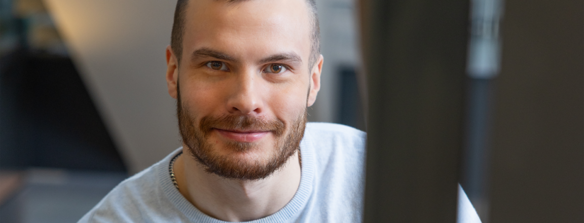 Mies, jolla on lyhyet tummat hiukset, hymyilee kameralle. Hänellä on yllään vaalea paita ja seisoo sisätiloissa nojaten porraskaiteeseen.