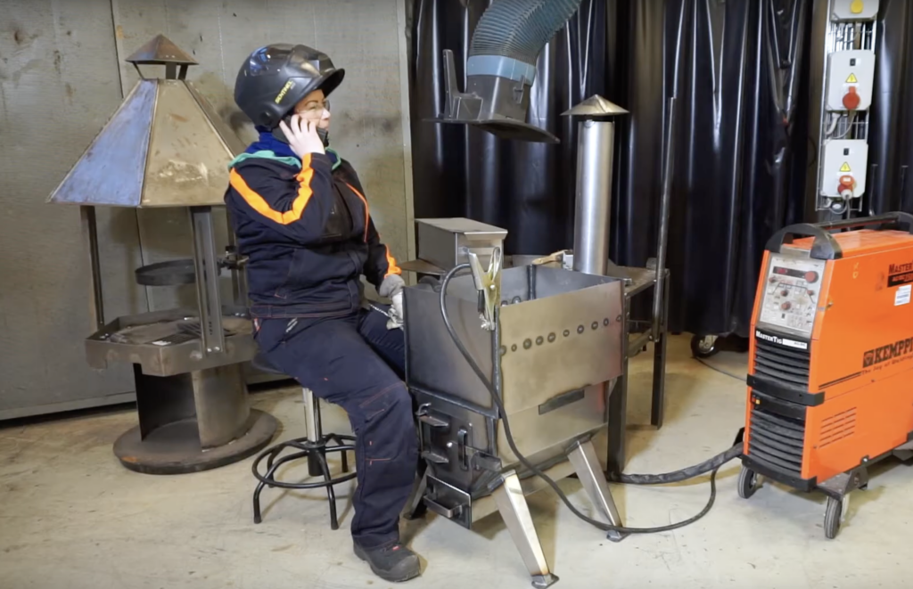 Henkilö istuu hitsauspajassa hitsauslaite vierellään, puhuu puhelimeen ja käyttää hitsauskypärää. Taustalla on metallinen grillirakenne ja sähköpisteitä.