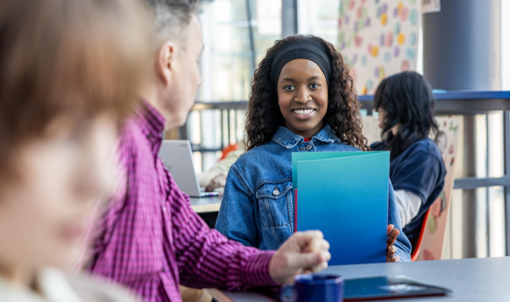 Iloinen nainen pitelee sinivihreää kansiota ja katsoo kameraan kahvilassa. Hänellä on yllään farkkutakki ja päänauha. Etualalla on toinen henkilö, joka kääntyy häntä kohti.