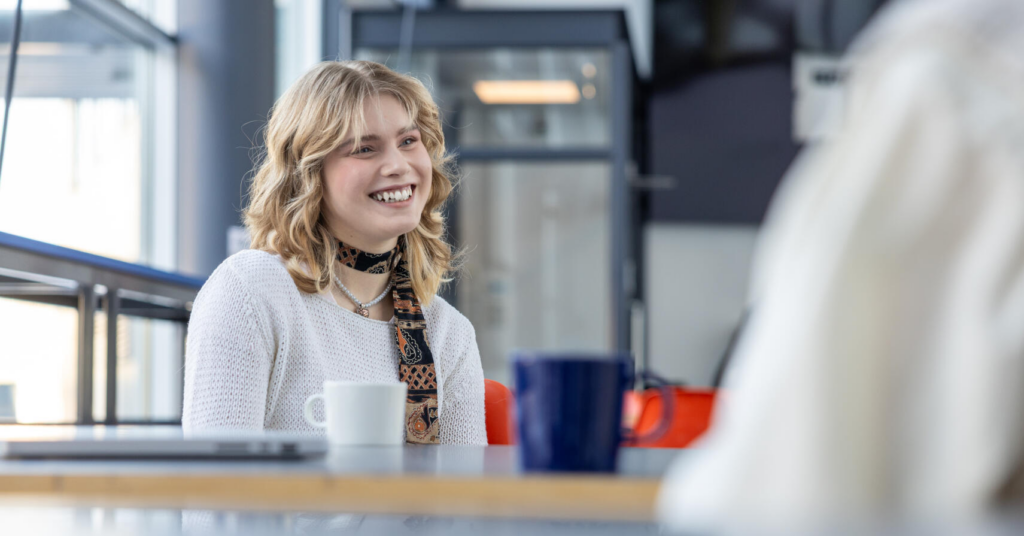 Nainen hymyilee kahvilassa, jossa on pöydällä kaksi mukia ja kannettava tietokone. Hänellä on päällään valkoinen paita ja kaulassa huivi. Taustalla näkyy ikkunat ja modernia sisustusta.