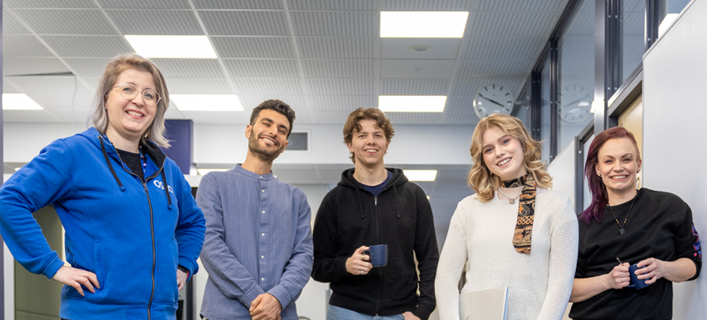 Viisi henkilöä seisoo sisätiloissa, hymyilee ja katsoo kameran suuntaan. Heillä on rento vaatetus, ja kaksi heistä pitää kahvikuppia. Taustalla näkyy toimiston katto ja kello seinällä.