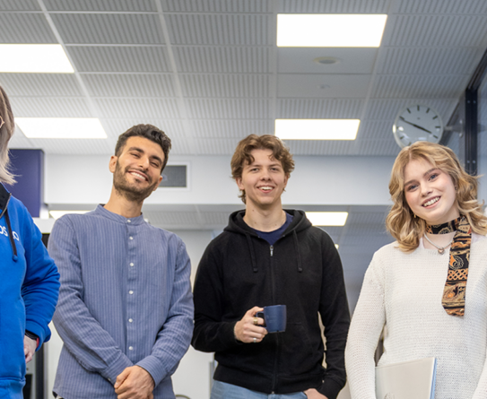 Viisi henkilöä seisoo sisätiloissa, hymyilee ja katsoo kameran suuntaan. Heillä on rento vaatetus, ja kaksi heistä pitää kahvikuppia. Taustalla näkyy toimiston katto ja kello seinällä.