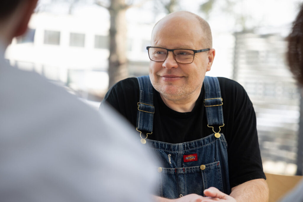 Mies hymyilee ja katsoo toista henkilöä. Hänellä on silmälasit ja yllään farkkuhaalarit ja musta paita. Tausta on epäselvä, mutta siinä on rakennuksen ikkunoita ja puun oksia.