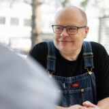 Mies hymyilee ja katsoo toista henkilöä. Hänellä on silmälasit ja yllään farkkuhaalarit ja musta paita. Tausta on epäselvä, mutta siinä on rakennuksen ikkunoita ja puun oksia.