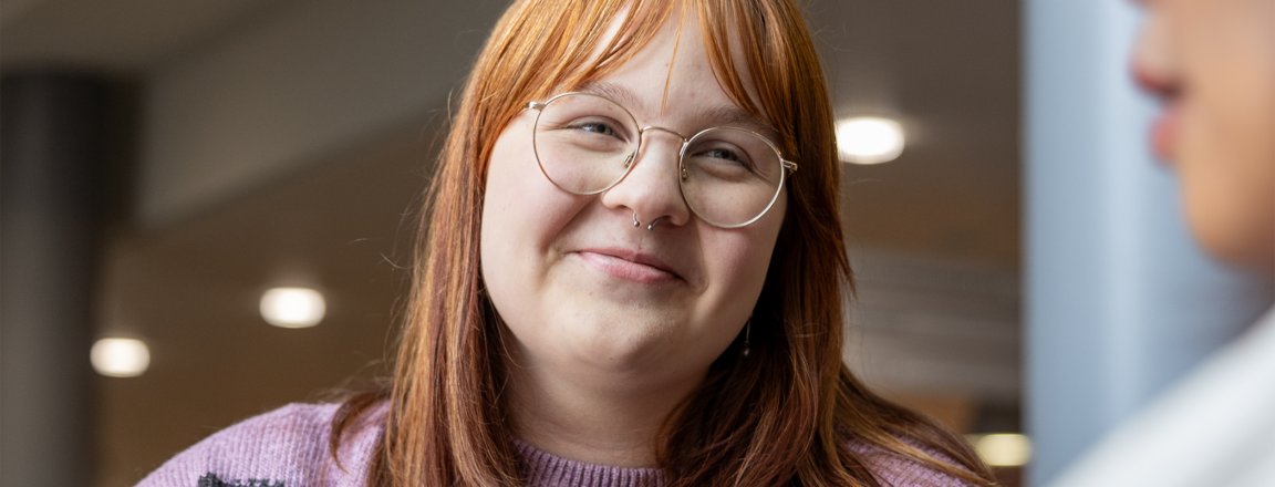 Henkilö, jolla on punertava tukka ja silmälasit, hymyilee sisätiloissa. Hänellä on yllään raidallinen villapaita. Toinen henkilö näkyy etualalla sivuprofiilista.