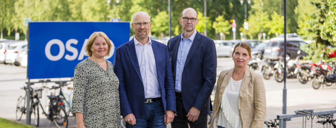 Neljä johtajaa seisovat OSAO-kyltin edessä koulun pihalla. Taustalla autoja ja polkupyöriä.