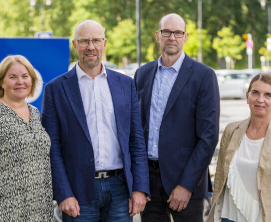 Neljä johtajaa seisovat OSAO-kyltin edessä koulun pihalla. Taustalla autoja ja polkupyöriä.