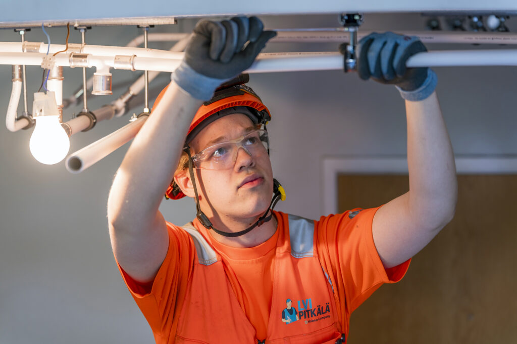 Putkiasentaja asentaa putkia kattoon sisätiloissa. Hänellä on suojalasit, kypärä ja oranssi työtakki."