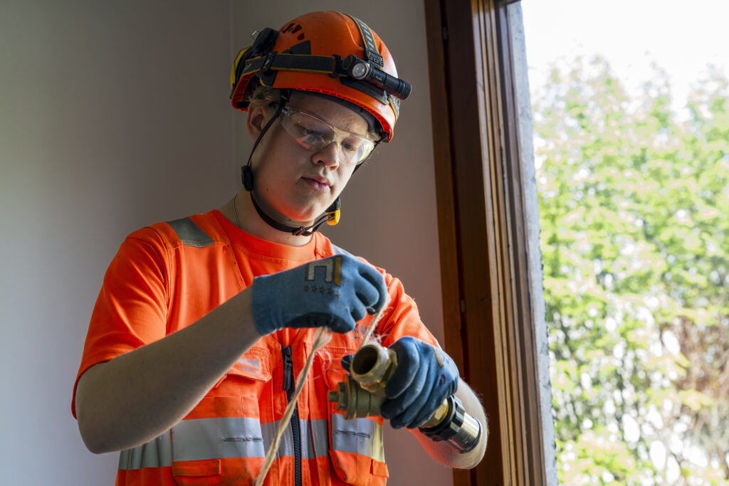 Rakennustyöntekijä pukeutuneena oranssiin huomiovaatteeseen ja kypärään, työskentelee putken parissa sisätiloissa. Hänellä on päässään suojalasit ja käsissään suojakäsineet.