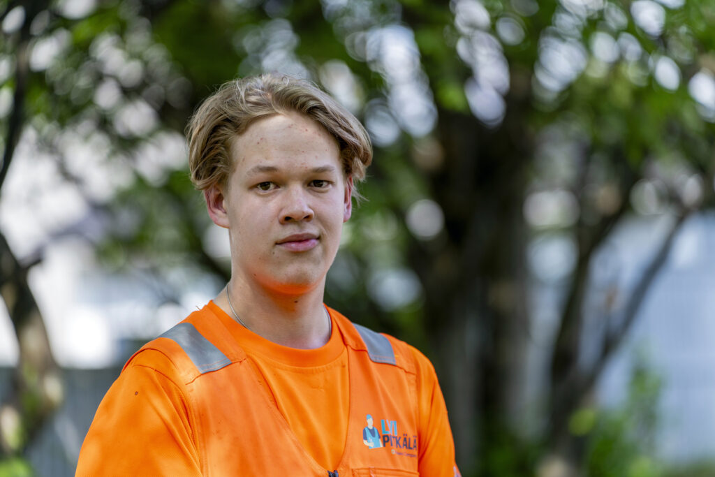 Nuori mies oranssissa työpaidassa seisoo ulkona puiden edessä. Hän katsoo suoraan kameraan.