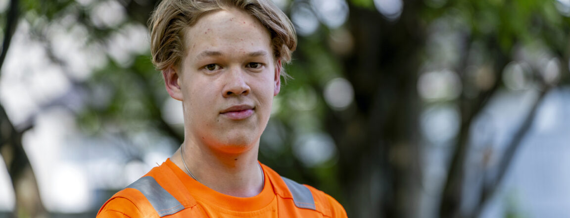 Nuori mies oranssissa työpaidassa seisoo ulkona puiden edessä. Hän katsoo suoraan kameraan.
