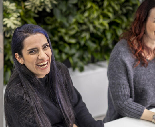 Kuvassa kaksi naista istuu pöydän ääressä ja nauraa. He ovat sisätiloissa, ja taustalla näkyy vihreitä kasveja. Molemmilla on pitkät hiukset, toinen on tumma- ja toinen punahiuksinen. Heillä on yllään neulepuserot.