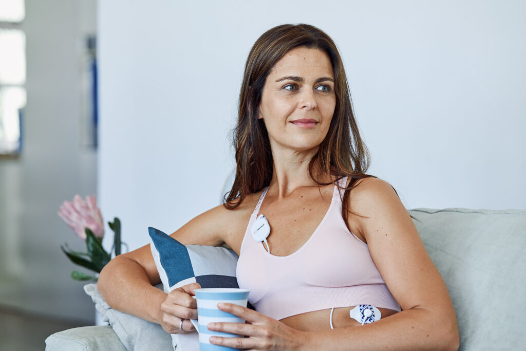 Nainen istuu sohvalla vaaleanpunaisessa urheilurintaliivissä, hymyillen hieman ja pitäen mukia kädessään. Hänellä on rintakehässään lääketieteellinen laite, ja hänen vieressään on sininen tyyny. Taustalla näkyy vaaleita kukkia maljakossa.