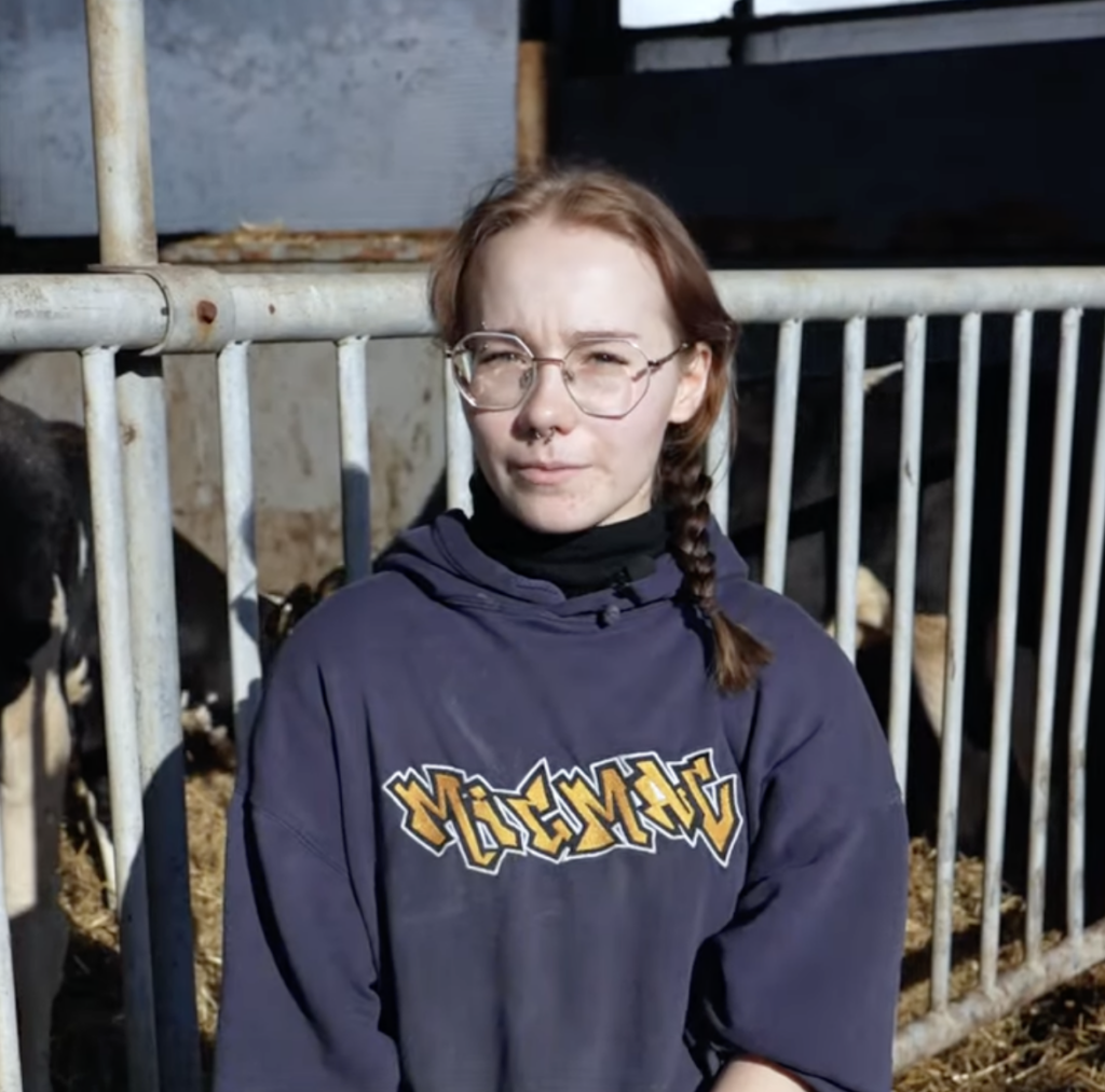 Henkilö navetassa aidan vieressä, yllään violetti huppari ja silmälasit. Taustalla näkyy lehmä.