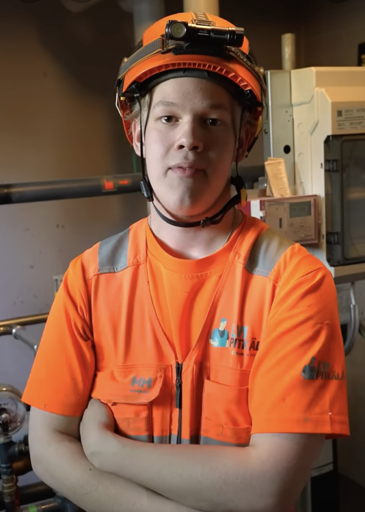 Henkilö pukeutunut kirkkaan oranssiin työasuun ja kypärään seisoo teollisessa ympäristössä, kädet ristissä rinnalla.