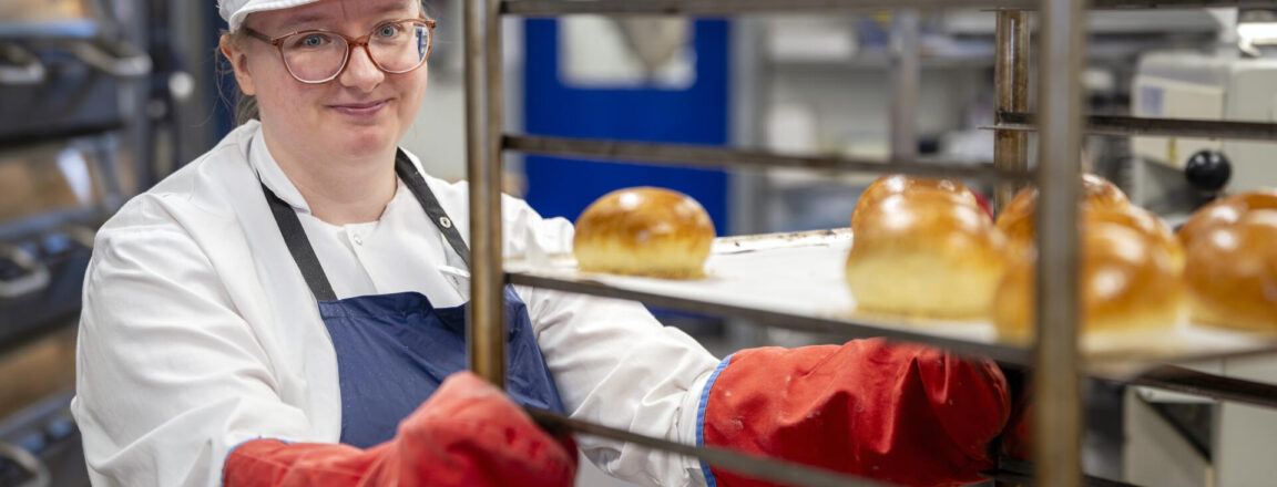 Leipuri siirtää uunituoreita leivonaisia metallisella kärryllä. Hänellä on yllään valkoinen lippalakki, esiliina ja punaiset uuninhanskat.