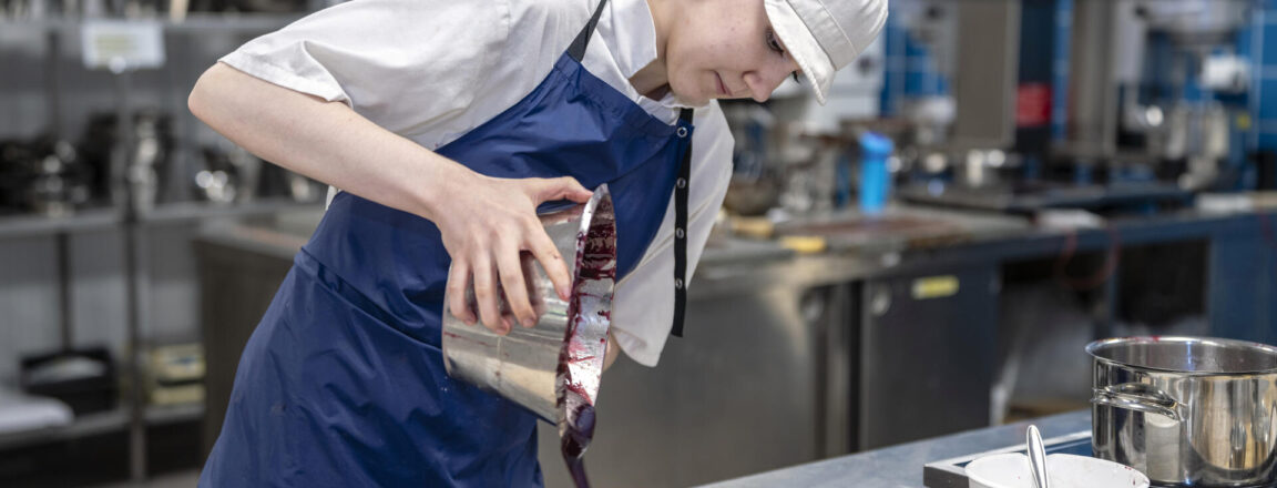 Kondiittori kaataa kastiketta kakun päälle keittiössä, jossa on siniset kaakeliseinät ja ruostumattomasta teräksestä valmistetut laitteet. Hänellä on valkoinen työasu ja esiliina.