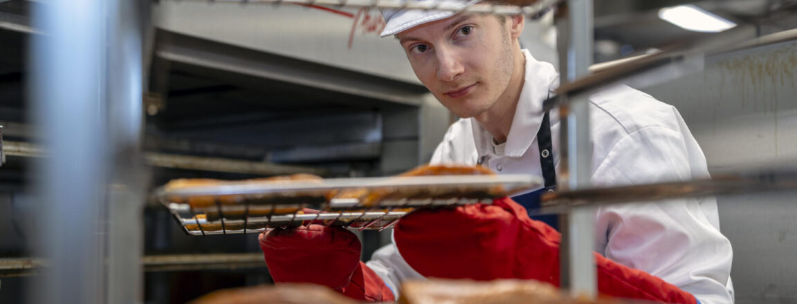 Lihatuotteiden valmistaja käyttää punaisia suojakäsineitä ja ottaa savustettua kalaa pois uunista keittiössä.