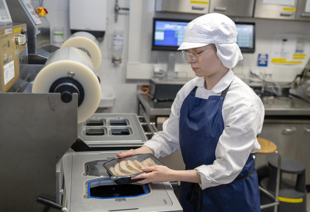 Henkilö pakkaamassa siivutettua lihaa muovirasiaan tehtaassa, yllään valkoinen suojahattu ja sininen esiliina.