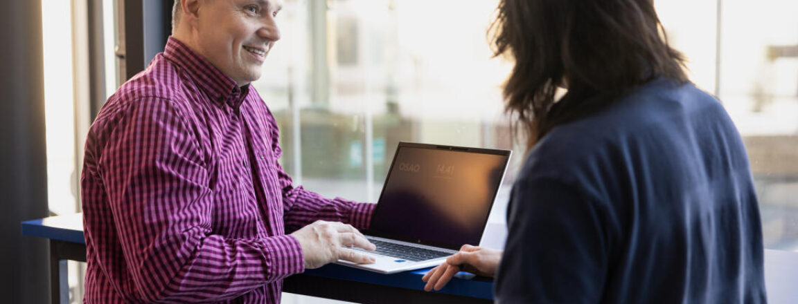 Kaksi henkilöä seisoo ikkunan ääressä, toinen heistä katsoo kannettavaa tietokonetta, joka on avoinna heidän välissään. Henkilö vasemmalla puolella on pukeutunut punaraitaisiin paitoihin ja hymyilee.