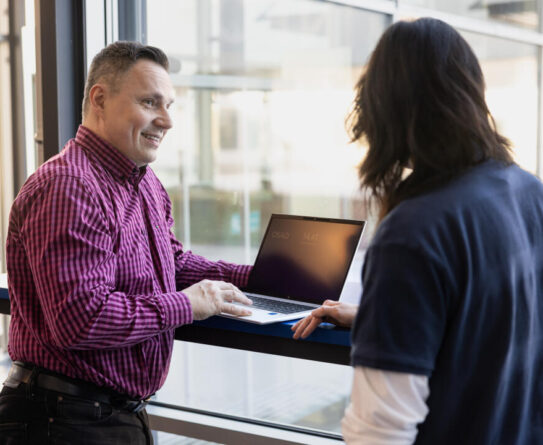 Kaksi henkilöä seisoo ikkunan ääressä, toinen heistä katsoo kannettavaa tietokonetta, joka on avoinna heidän välissään. Henkilö vasemmalla puolella on pukeutunut punaraitaisiin paitoihin ja hymyilee.