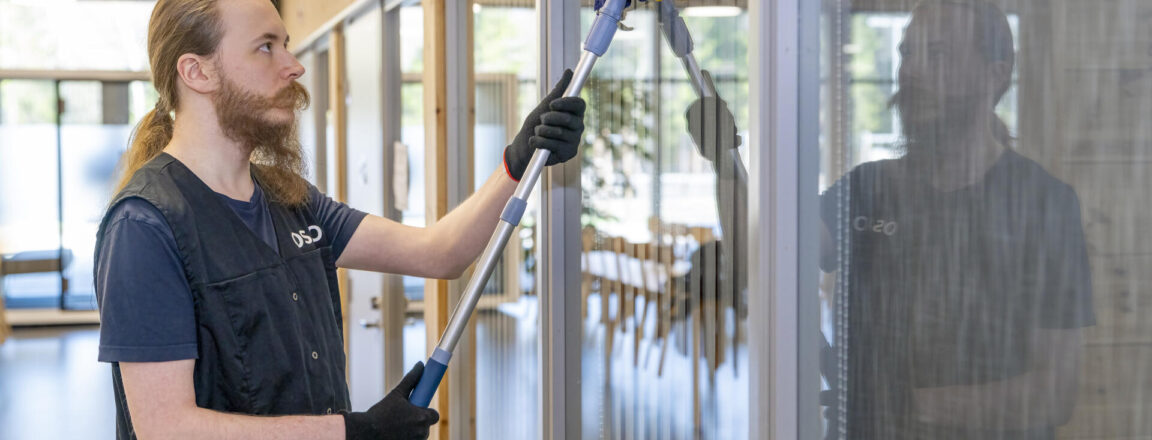 Henkilö puhdistaa lasiovea tai -ikkuvaa sisätiloissa. Hänellä on päällään musta liivi ja käsineet.