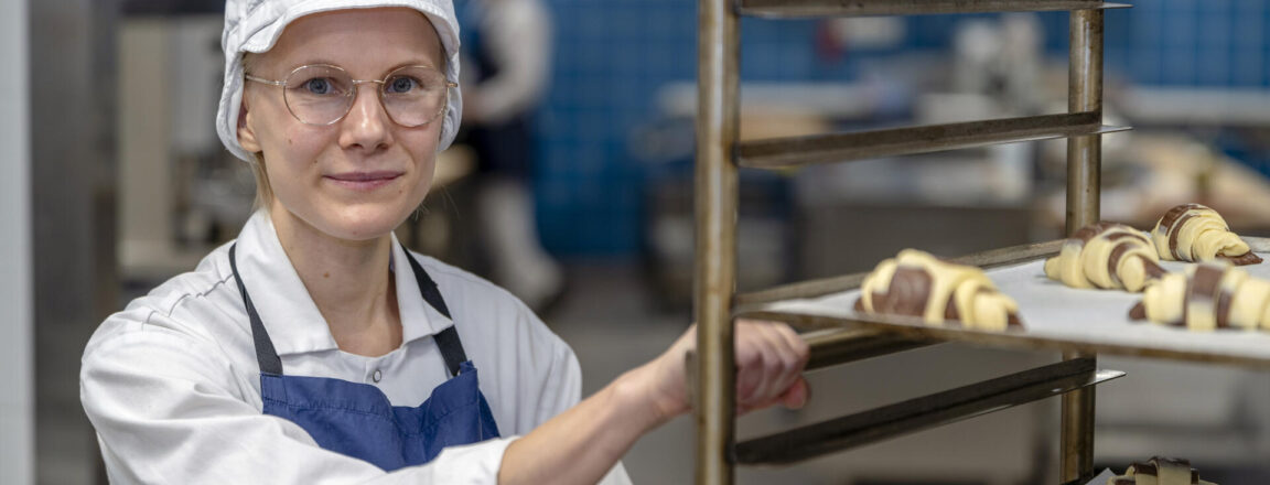 Leipomotyöntekijä, jolla on valkoinen huivi ja sininen esiliina, seisoo metallisten leipäritilöiden vieressä. Telineillä on raakoja leivonnaisia menossa uuniin.