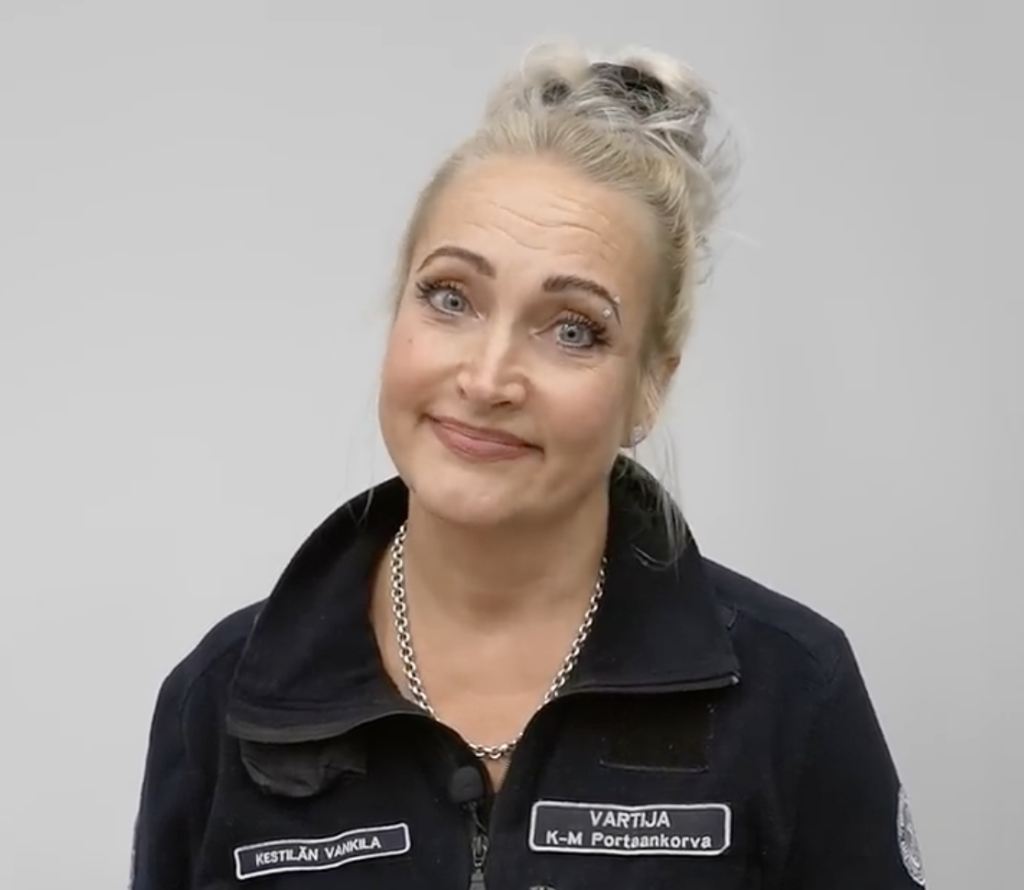 Vaaleahiuksinen nainen hymyilee ja katsoo kameraan. Hänellä on musta takki, jossa on nimikyltit. Tausta on yksivärinen harmaa.