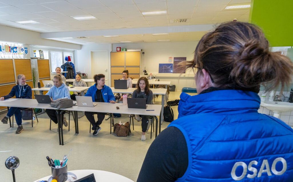 Opettaja seisoo luokan edessä puhumassa opiskelijoille, jotka istuvat pulpeteilla kannettavien tietokoneiden kanssa.