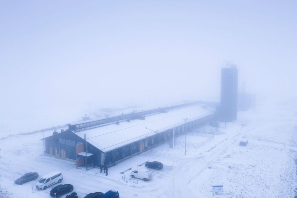 Luminen maisema, jossa on suuri navettarakennus ja useita autoja pysäköitynä etualalla. Taustalla näkyy sumua, peltoja ja korkeita rakenteita.