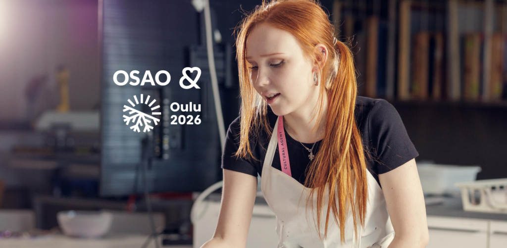 Nuori nainen seisoo tekstiilialan luokassa päällään esiliina ja kaulassa kaulanauha, jossa lukee Cultural Agent. Hänen vieressään on OSAO ja Oulu2026 logot. Hänellä on pitkät punaiset hiukset ja hän katsoo alaspäin keskittyneenä johonkin edessään olevassa työssä.