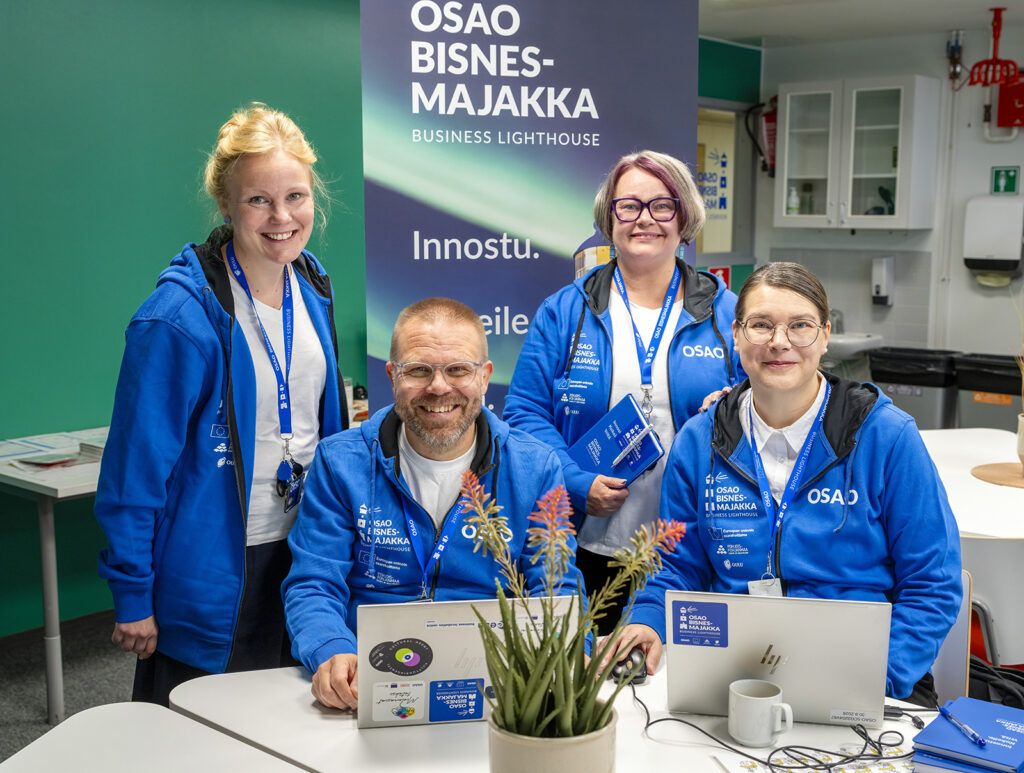 Neljä henkilöä seisoo ja istuu pöydän ääressä, kaikki pukeutuneina sinisiin OSAO-huppareihin. He hymyilevät kameralle. Pöydällä on kannettava tietokone ja kasvi. Taustalla on roll up, jossa lukee "OSAO Bisnesmajakka Business Lighthouse" ja "Innostu."