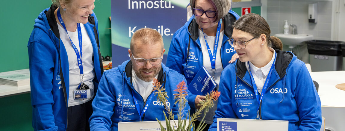 Neljä henkilöä sinisissä huppareissa työskentelee yhdessä kannettavien tietokoneiden ääressä sisätiloissa. Taustalla on Bisnesmajakka Business Lighthouse -roll up, jossa lukee: "Innostu. Kokeile." Keskellä pöytää on kasvi. Kaikilla henkilöillä on nimikyltit kaulassaan.