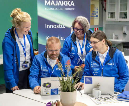 Neljä henkilöä sinisissä huppareissa työskentelee yhdessä kannettavien tietokoneiden ääressä sisätiloissa. Taustalla on Bisnesmajakka Business Lighthouse -roll up, jossa lukee: "Innostu. Kokeile." Keskellä pöytää on kasvi. Kaikilla henkilöillä on nimikyltit kaulassaan.