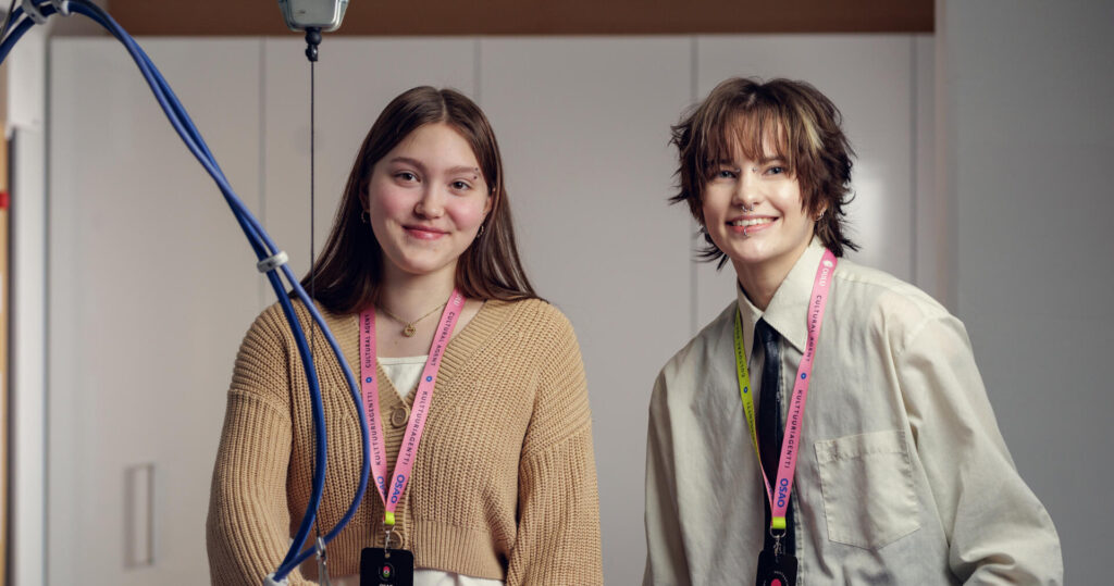 Kaksi tekstiili- ja muotialan opiskelijaa poseeraa hymyillen kameralle silityspöydän ääressä. Molemmilla on kaulassaan värikkäät Kulttuuriagentti-kaulanauhat.