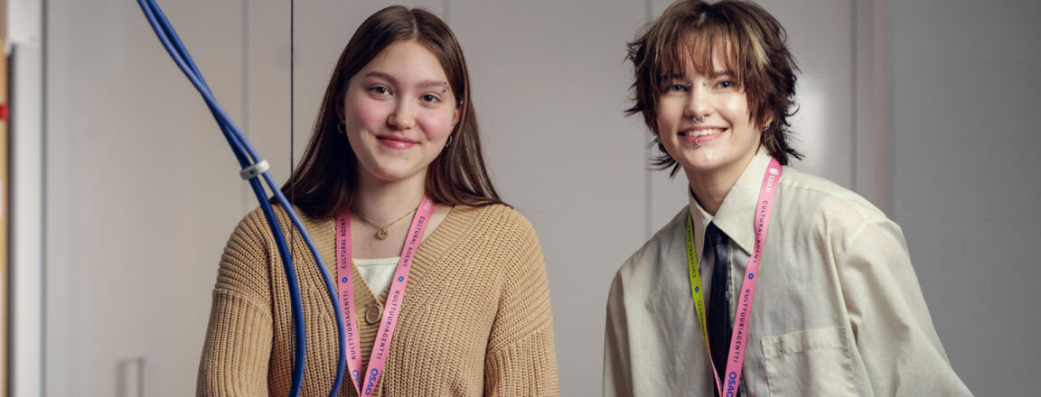 Kaksi tekstiili- ja muotialan opiskelijaa poseeraa hymyillen kameralle silityspöydän ääressä. Molemmilla on kaulassaan värikkäät Kulttuuriagentti-kaulanauhat.