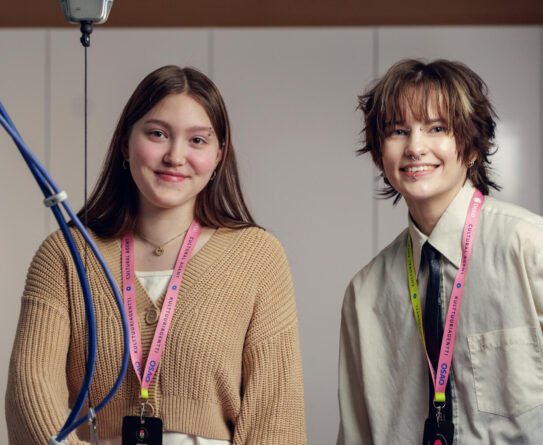 Kaksi tekstiili- ja muotialan opiskelijaa poseeraa hymyillen kameralle silityspöydän ääressä. Molemmilla on kaulassaan värikkäät Kulttuuriagentti-kaulanauhat.