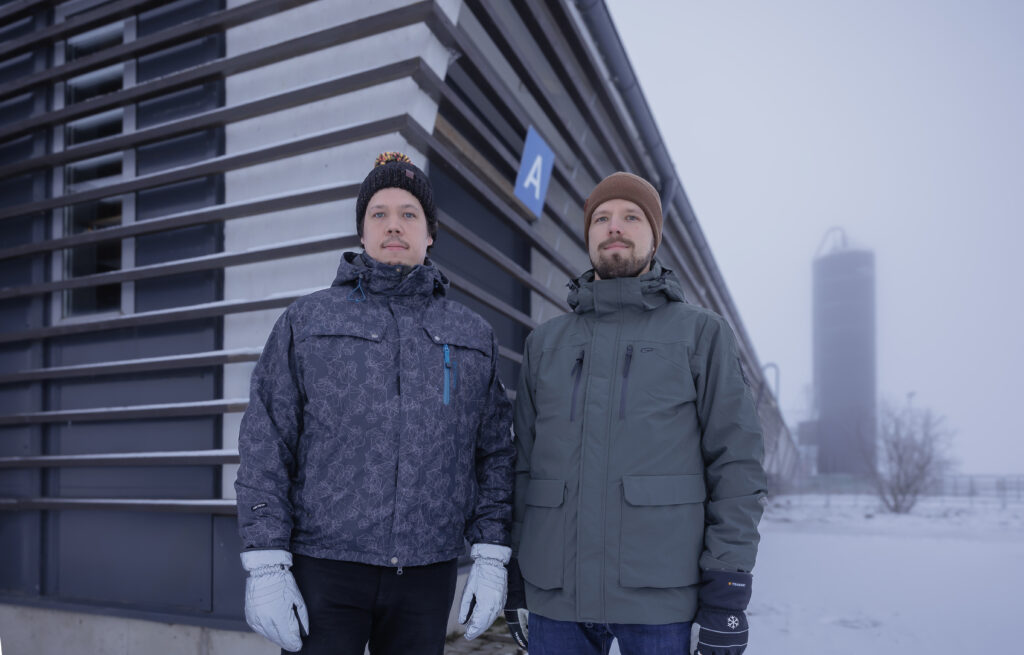 Kaksi miestä seisoo ulkona talvitakeissa ja pipoissa lumisessa ympäristössä. Heidän takanaan on moderni rakennus ja kauempana taustalla näkyy siilo.