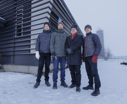 Neljän miehen ryhmä seisoo lumisessa ympäristössä navetan edessä. He ovat pukeutuneet talvivaatteisiin, kuten takkeihin, hanskoihin ja pipoihin. Taustalla näkyy sumuinen peltomaisema ja toinen rakennus.