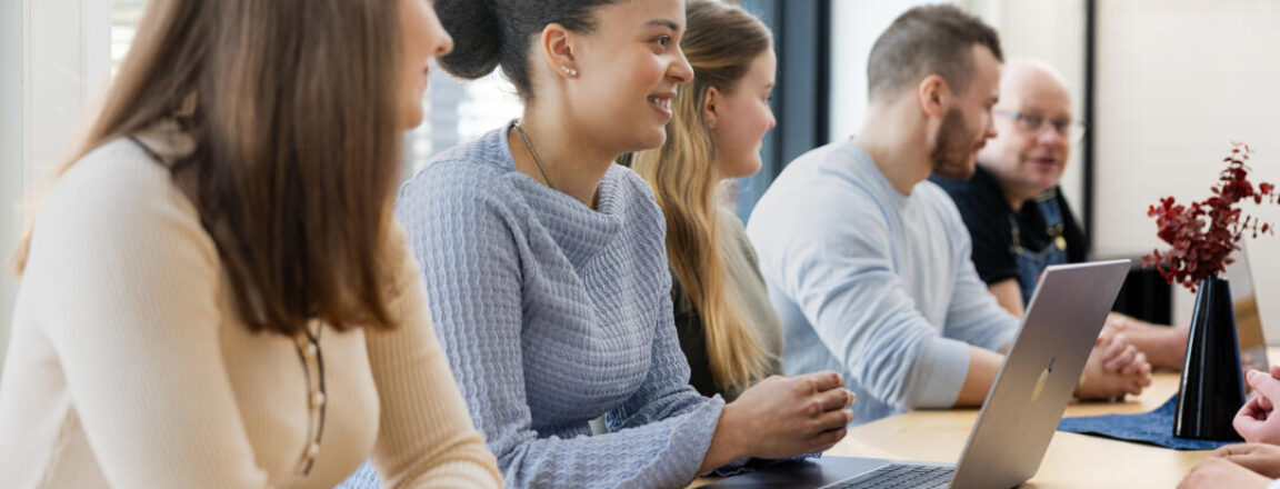 Ryhmä henkilöitä istuu pöydän ääressä kokouksessa. Heillä on kannettava tietokone edessään ja he keskustelevat keskenään. Ikkuna taustalla tuo luonnonvaloa.