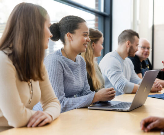 Ryhmä henkilöitä istuu pöydän ääressä kokouksessa. Heillä on kannettava tietokone edessään ja he keskustelevat keskenään. Ikkuna taustalla tuo luonnonvaloa.