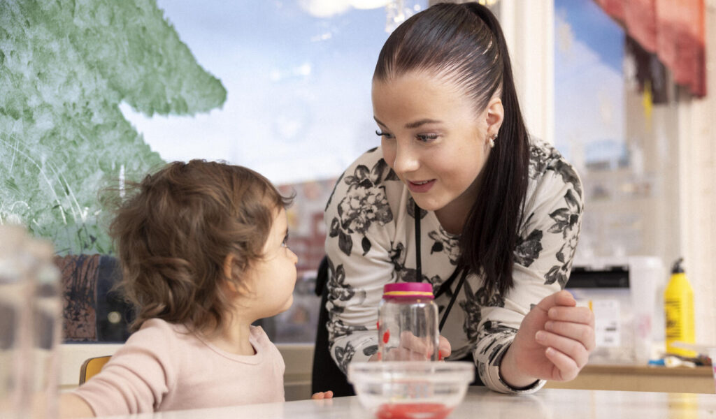 Aikuinen nainen hymyilee pienelle lapselle, joka istuu pöydän ääressä. He ovat sisätiloissa vaaleassa huoneessa, jonka taustalla on ikkunasta näkyvä puu. Pöydällä on leluja ja astioita. Nainen ja lapsi näyttävät keskustelevan keskenään ystävällisesti.