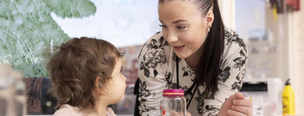 Aikuinen nainen hymyilee pienelle lapselle, joka istuu pöydän ääressä. He ovat sisätiloissa vaaleassa huoneessa, jonka taustalla on ikkunasta näkyvä puu. Pöydällä on leluja ja astioita. Nainen ja lapsi näyttävät keskustelevan keskenään ystävällisesti.