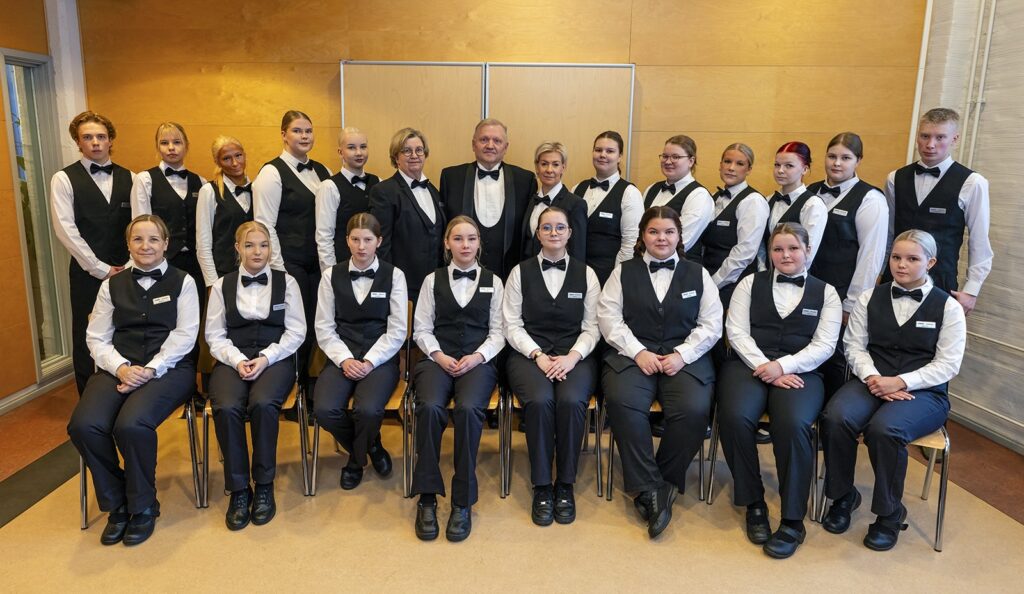 Ryhmä nuoria pukeutuneena tarjoilijan asuihin istuu ja seisoo rivissä sisätiloissa. Heillä on valkoiset paidat, mustat liivit ja solmiot. Taustalla on vaaleanruskea seinä.