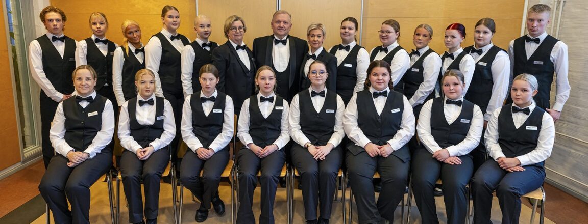 Ryhmä nuoria pukeutuneena tarjoilijan asuihin istuu ja seisoo rivissä sisätiloissa. Heillä on valkoiset paidat, mustat liivit ja solmiot. Taustalla on vaaleanruskea seinä.