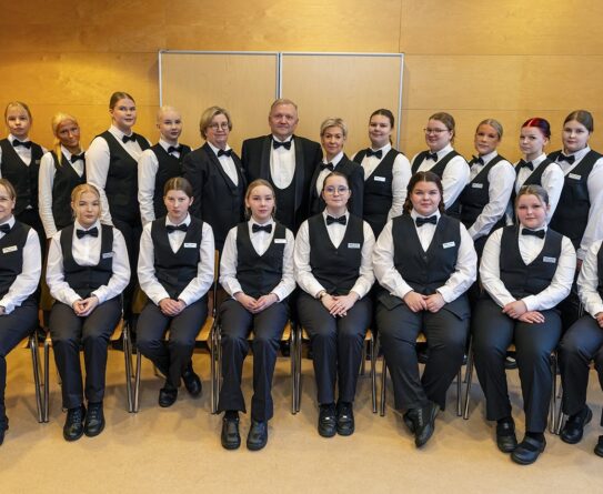 Ryhmä nuoria pukeutuneena tarjoilijan asuihin istuu ja seisoo rivissä sisätiloissa. Heillä on valkoiset paidat, mustat liivit ja solmiot. Taustalla on vaaleanruskea seinä.