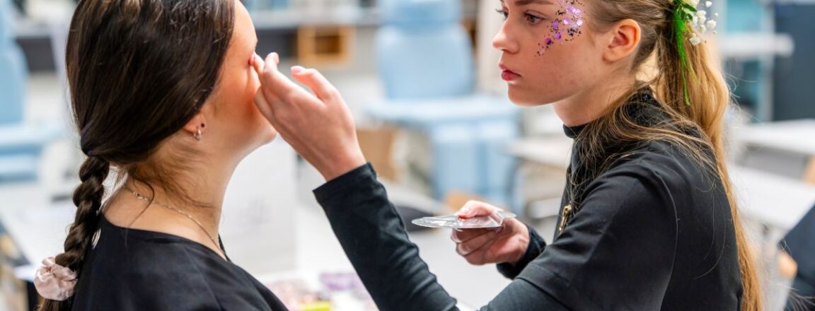 Kaksi nuorta naista kauneushoitolassa, toinen tekee meikkiä toiselle. Vasemmalla oleva nainen istuu, kun oikealla oleva nainen levittää meikkiä. Oikealla oleva nainen on koristeltu kasvoihin kiinnitetyillä koristeilla ja kukilla hiuksissaan.