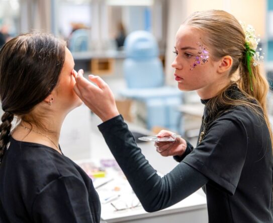 Kaksi nuorta naista kauneushoitolassa, toinen tekee meikkiä toiselle. Vasemmalla oleva nainen istuu, kun oikealla oleva nainen levittää meikkiä. Oikealla oleva nainen on koristeltu kasvoihin kiinnitetyillä koristeilla ja kukilla hiuksissaan.